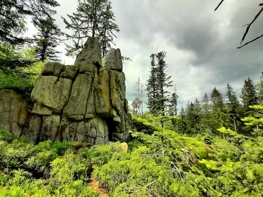 Massive ancient rock formations covered in moss and evergreen trees under a cloudy sky at the Heathen Stones near Oberlichtbuchet