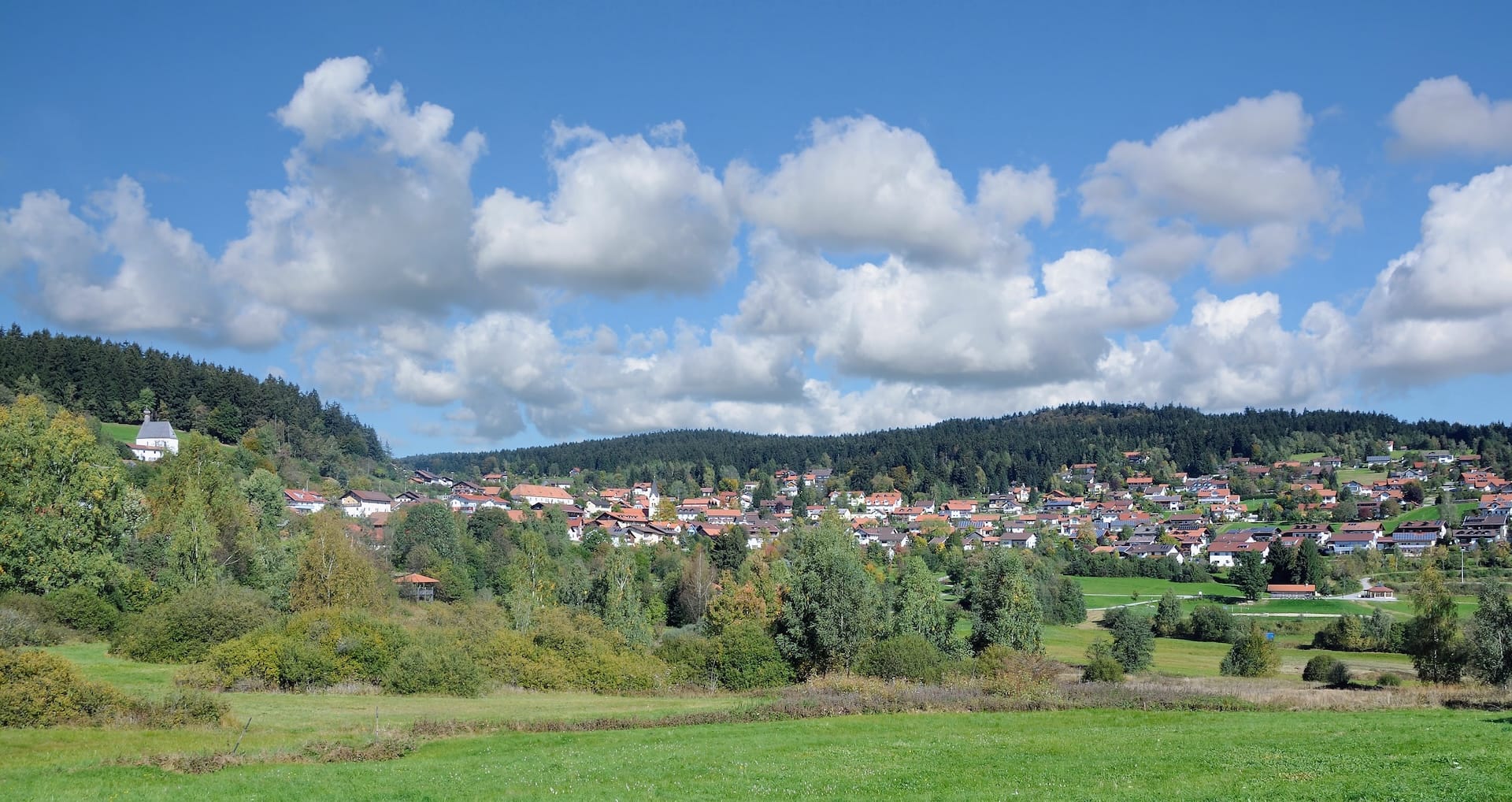Wide panoramic view of the picturesque mountain village of Sankt Englmar nestled in the green hills and dense pine forests of the Bavarian Forest, under a bright blue sky with fluffy white clouds.