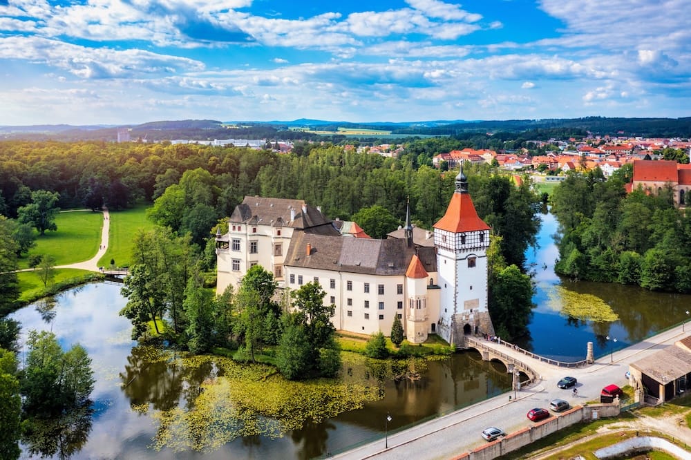 High-angle aerial view of Blatná Castle in South Bohemia, showing the white-walled water castle and its red-roofed tower surrounded by a wide moat, with a stone bridge and the green trees of the landscape park