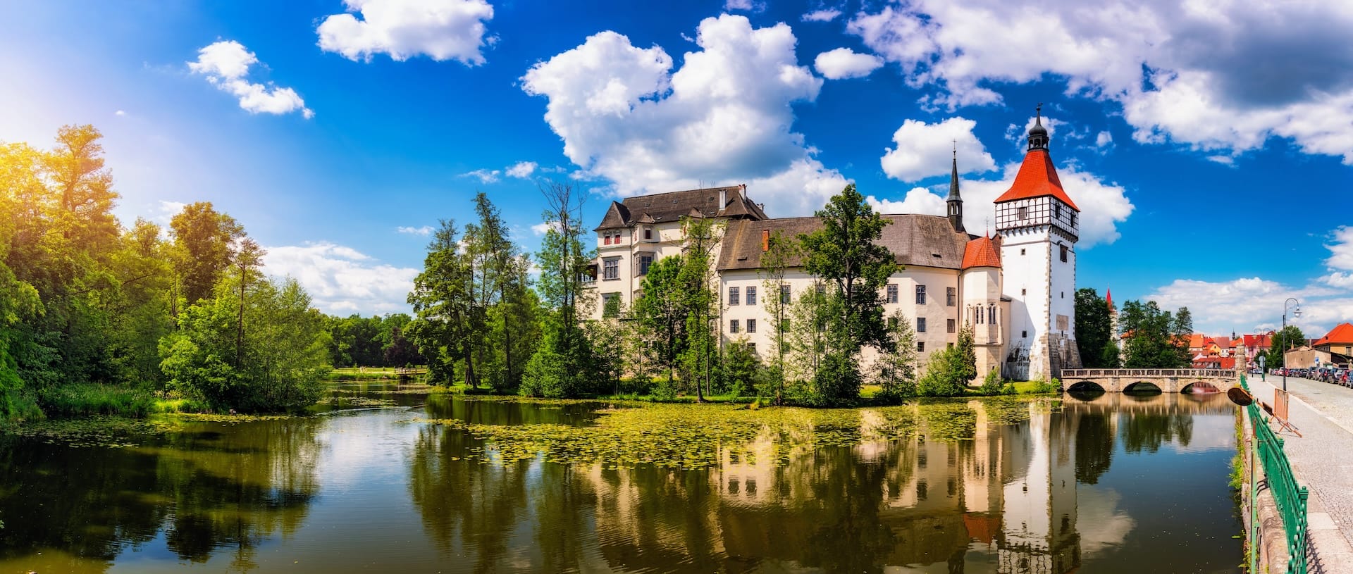 Panorama of the historic Blatná water castle with its red-roofed tower reflecting in the calm surrounding lake under a bright blue sky in South Bohemia