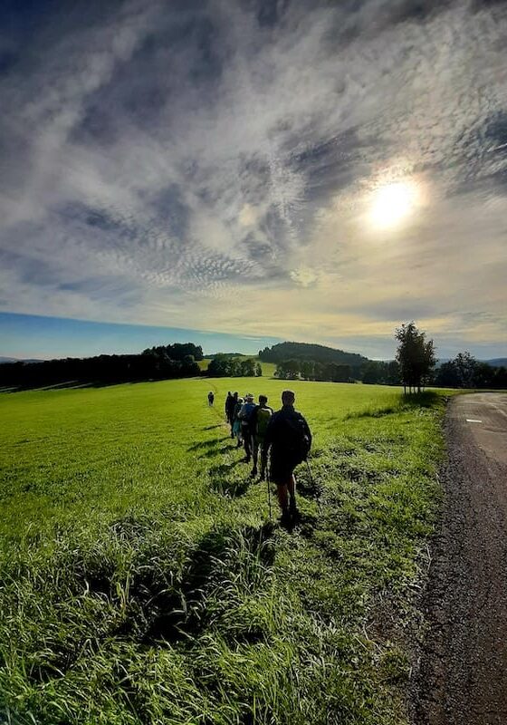 A line of hikers walking through an open green field in a quiet, lesser-known European landscape under a vast sky filled with dramatic clouds and bright sun
